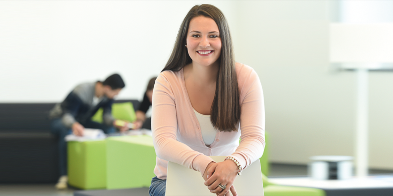 Junge Frau lehnt an Stuhl. Im Hintergrund arbeiten Studierende in Sitzgruppen.