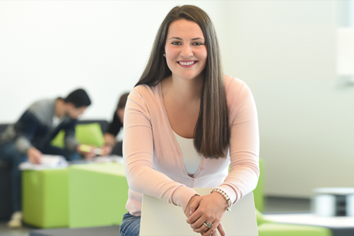 Junge Frau lehnt an Stuhl. Im Hintergrund arbeiten Studierende in Sitzgruppen.