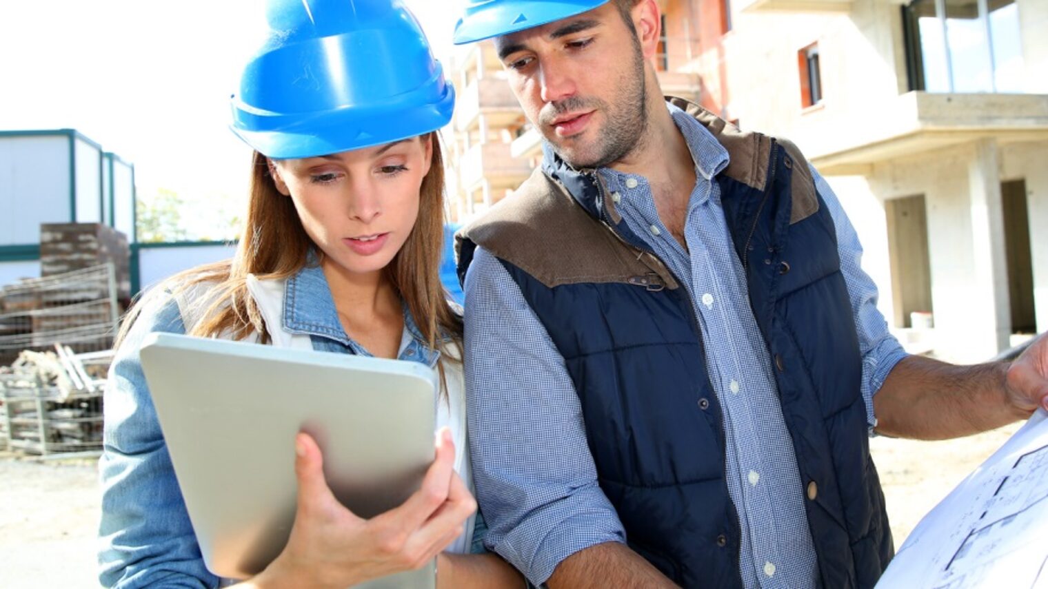 Eine Frau und ein Mann mit Schutzhelmen auf einer Baustelle. Sie hält ein Tablet in der Hand, er eine Bauzeichnung.