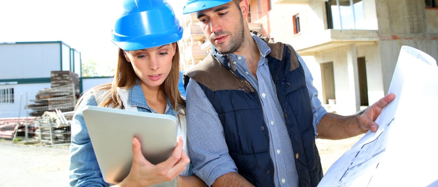 Eine Frau und ein Mann mit Schutzhelmen auf einer Baustelle. Sie hält ein Tablet in der Hand, er eine Bauzeichnung.