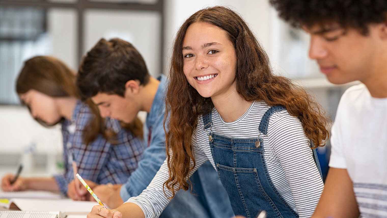 Schülerin sitzt mit ihren Klassenkameraden in einer Reihe und schaut in die Kamera.