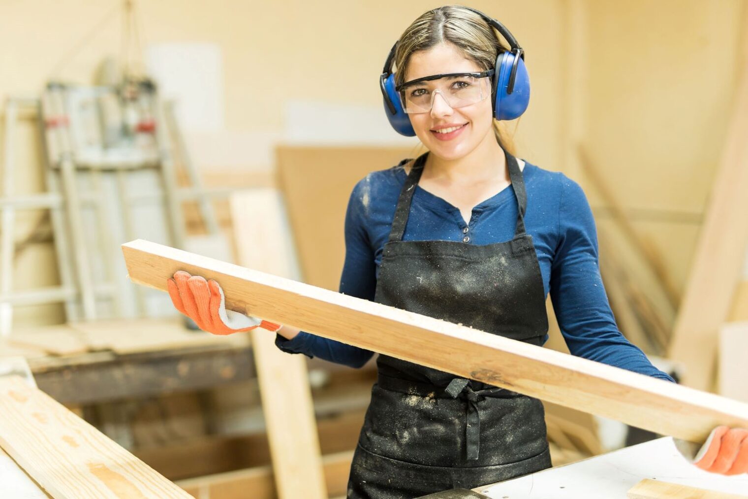 Junge Tischlerin in einer Werkstatt hält eine Holzplanke in den Händen und blickt in die Kamera.