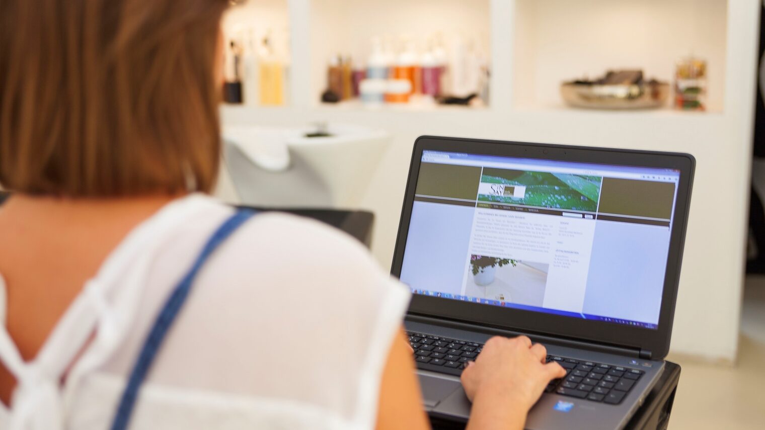 Rückenansicht einer Friseurinhaberin, die an ihrem Laptop arbeitet.