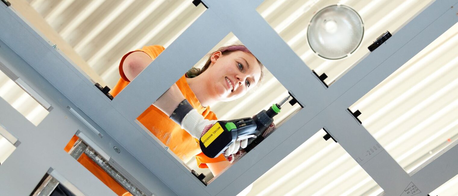 Eine Tischlerin, von unten fotografiert, arbeitet mit einer Bohrmaschine an einer Deckenkonstruktion.