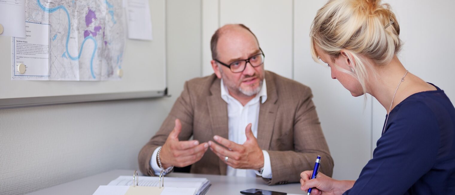 Beratungsgespräch zwischen einem Mann und einer Frau. Auf dem Tisch liegt ein geöffneter Ordner mit Unterlagen. Die Frau macht sich Notizen.