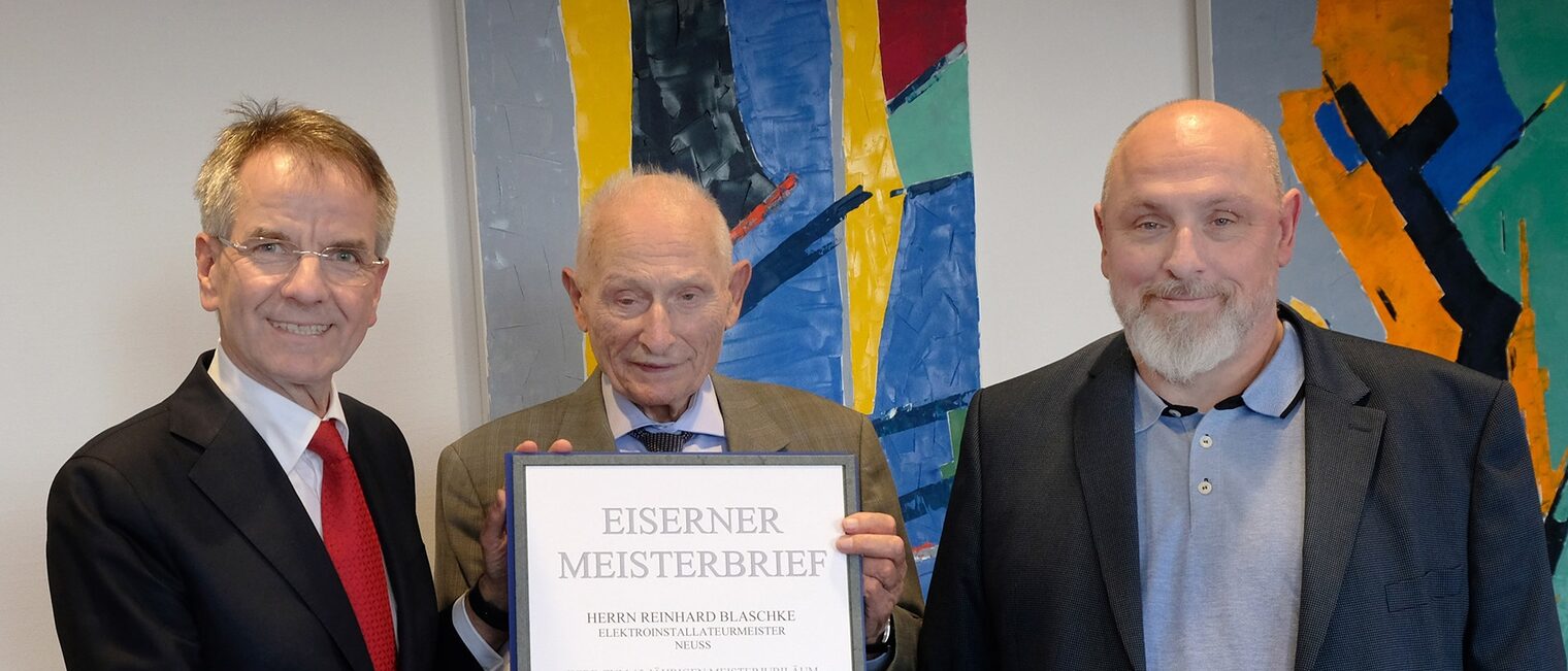 Reinhard Blaschke zeigt seinen Eisernen Meisterbrief. Er posiert mit zwei Gratulanten vor zwei bunten Bildern.